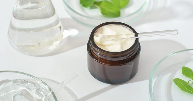 Brown jar of cream with a spatula surrounded by clear liquid and green leaves, representing sustainable cosmetics manufacturing.