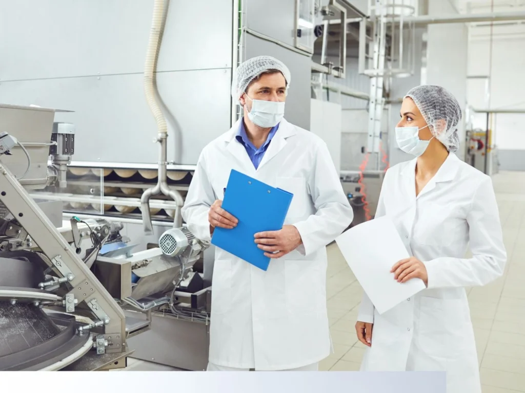 Two GMP-compliant professionals in lab coats and hairnets inspecting equipment in a clean manufacturing facility.