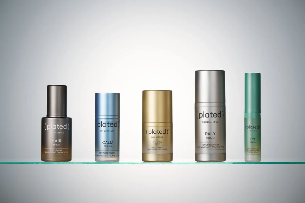 Five plated skincare serum bottles in different colors and sizes arranged on a glass shelf against a gray background.