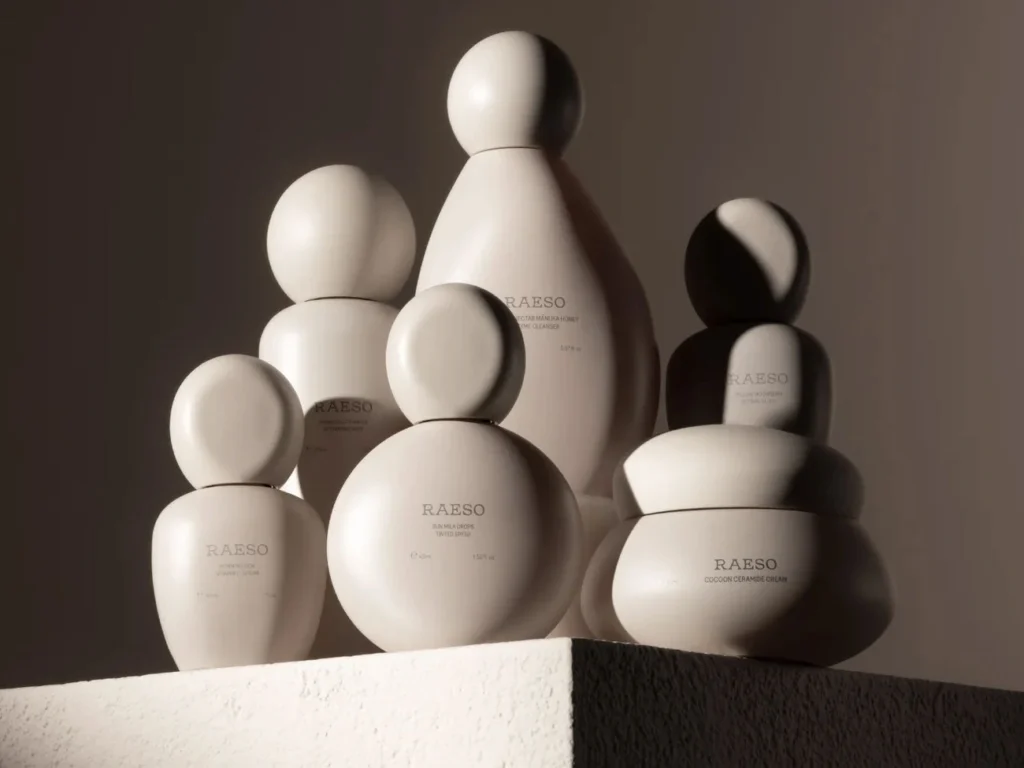 Six minimalist beige Raeso skincare product containers with round caps displayed on a textured pedestal with soft lighting.